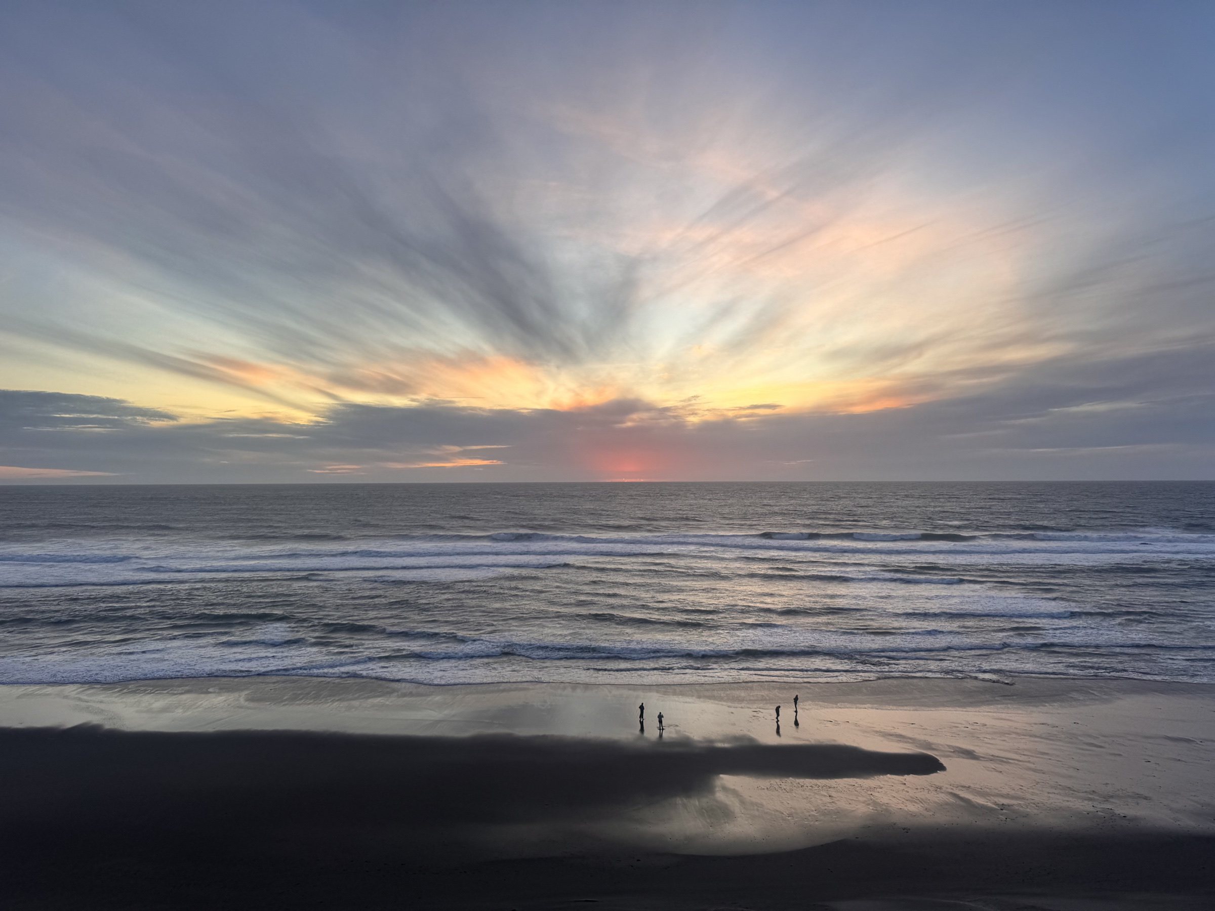 Sunset opens over the ocean as small figures stand along the shoreline.