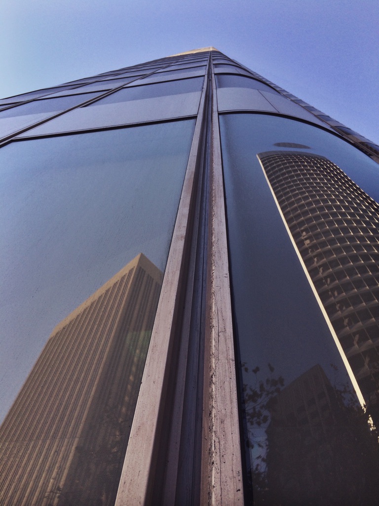 A glass building rises into the sky, reflecting neighboring towers.