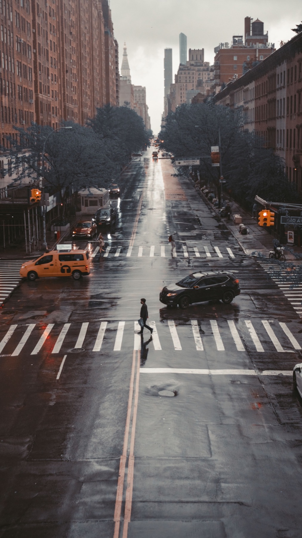 A person crosses a broad city intersection after rain while traffic moves around them.